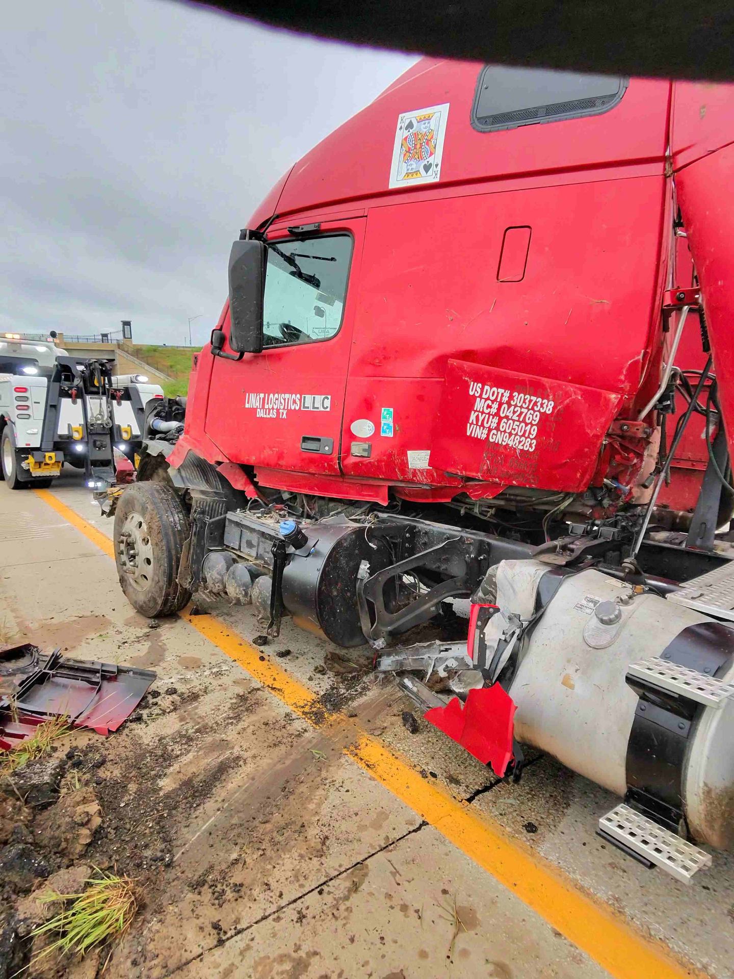 Titan Tow & Repair heavy-duty wrecker on-scene recovering a wrecked red Volvo VNL sleeper semi tractor from Linat Logistics (Dallas, TX) after a highway collision in the Kansas City metro — front cab and steer axle damaged, bumper and grille debris on the shoulder, Titan Class-8 heavy wrecker staged behind for hookup.